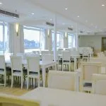 A bright, all-white cruise ship restaurant with large windows overlooking the Nile, featuring long dining tables set with white tablecloths and light beige/white upholstered chairs.