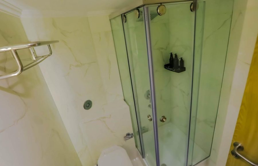 A modern cruise cabin bathroom with a corner glass-enclosed shower, a white toilet, and walls covered in light marble-look tiles with gold veining.