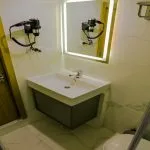 A modern cruise cabin bathroom sink area with a square basin, a vanity mirror with built-in LED lighting, a wall-mounted hairdryer, and white marble-look wall tiles.