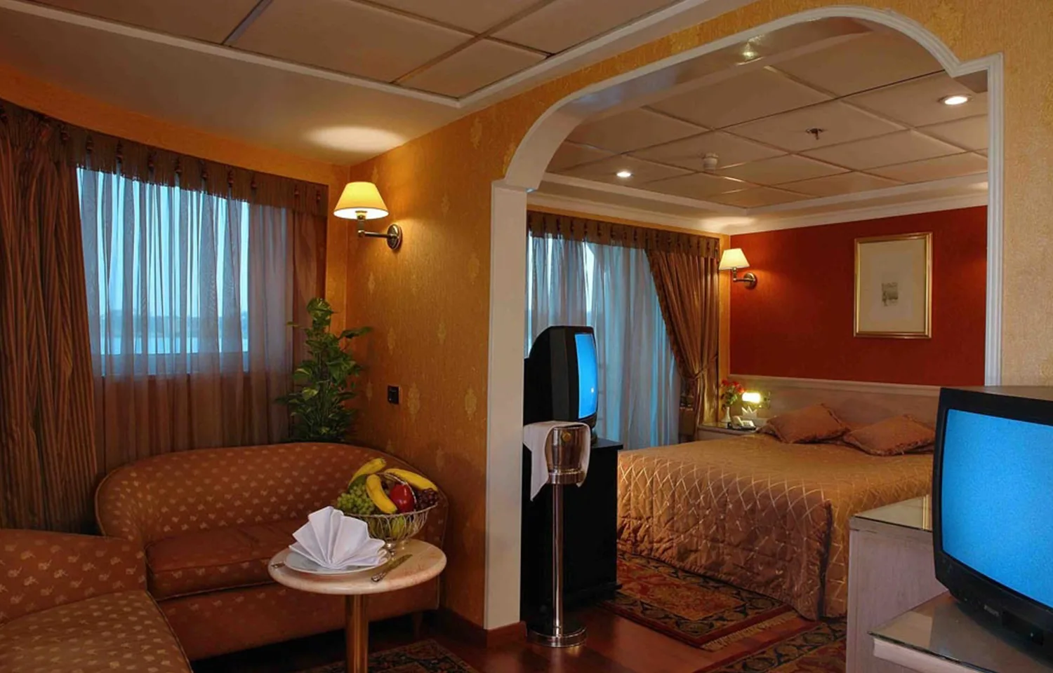 Interior of a Nile cruise suite or cabin divided by an archway into a sitting area and a bedroom, featuring warm orange and red colors.