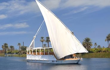 Nora Dahabiya Nile Cruise boat sailing on the water past palm trees.