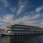 A long shot of the MS Nile Odyssey cruise ship moving on the water, featuring its white and green multiple-deck structure against a dramatic blue sky with sweeping clouds.