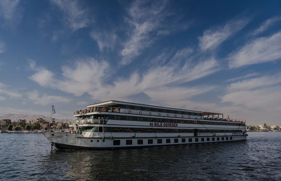 A long shot of the MS Nile Odyssey cruise ship moving on the water, featuring its white and green multiple-deck structure against a dramatic blue sky with sweeping clouds.