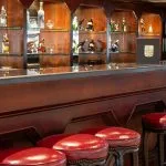 Close-up of the MS Odyssey bar counter, showing the dark wood paneling, red leather-topped stools, granite countertop, and illuminated glass shelves stocked with liquor.