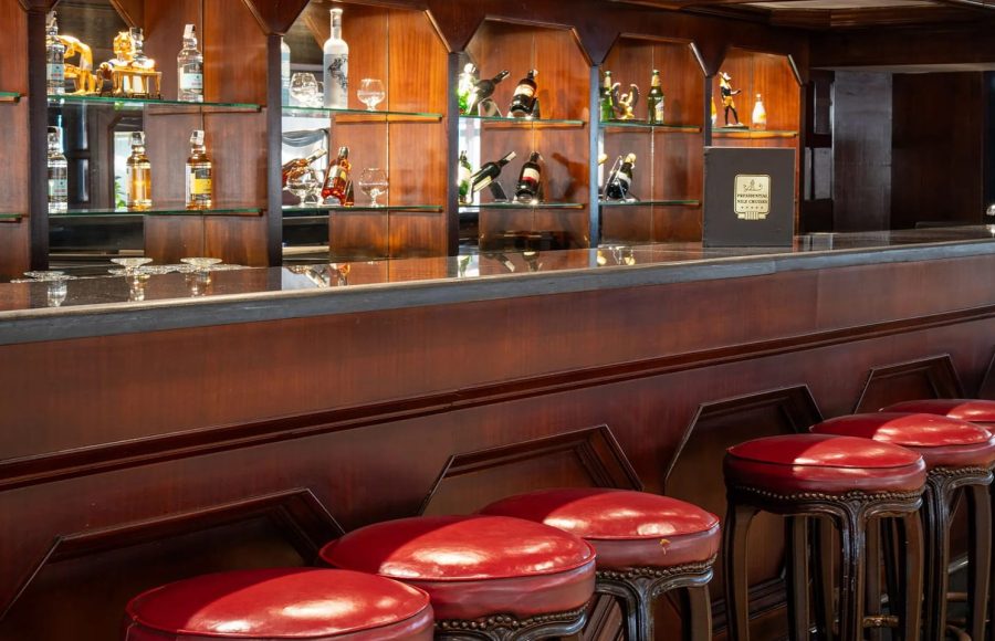 Close-up of the MS Odyssey bar counter, showing the dark wood paneling, red leather-topped stools, granite countertop, and illuminated glass shelves stocked with liquor.