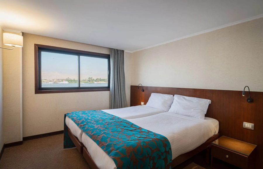 A comfortable, bright guest cabin on the cruise ship featuring two separate twin beds pushed together, a modern wooden headboard, and a large window with a view of the Nile.