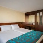 Interior view of a cruise ship cabin, showing the twin beds pushed together, a wooden headboard, and a built-in vanity unit with mirrors and storage.