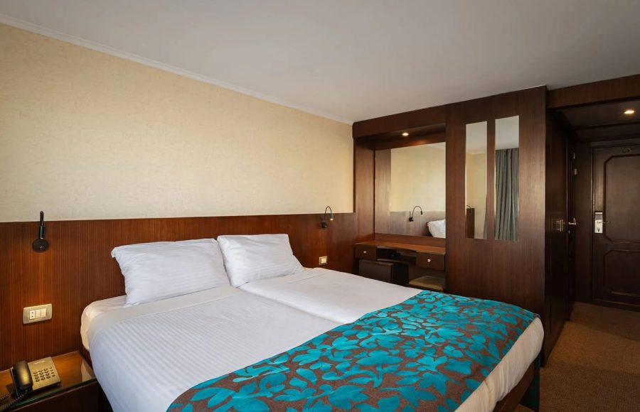 Interior view of a cruise ship cabin, showing the twin beds pushed together, a wooden headboard, and a built-in vanity unit with mirrors and storage.