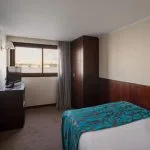 View of a single twin bed cabin on a cruise ship, showing the dark wood headboard, a television on a cabinet, a framed mirror, and a window with a view of the river.