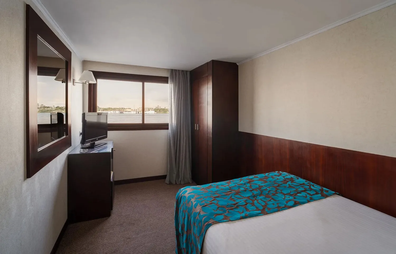 View of a single twin bed cabin on a cruise ship, showing the dark wood headboard, a television on a cabinet, a framed mirror, and a window with a view of the river.