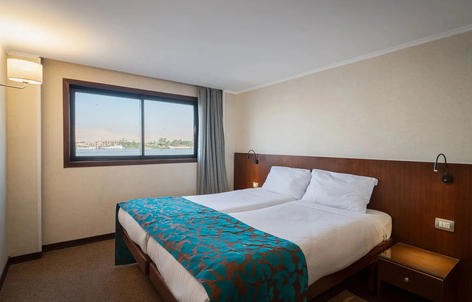 A comfortable, bright guest cabin on the cruise ship featuring two separate twin beds pushed together, a modern wooden headboard, and a large window with a view of the Nile.