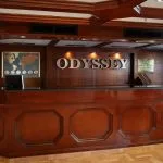 The classic, dark wood reception and check-in desk of the MS Odyssey cruise ship. The word "ODYSSEY" is mounted on the wall behind the desk.