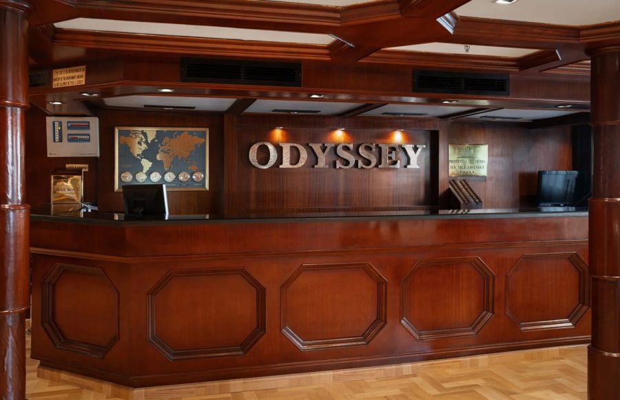 The classic, dark wood reception and check-in desk of the MS Odyssey cruise ship. The word "ODYSSEY" is mounted on the wall behind the desk.