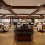 A wide-angle view of the large and elegant dining room on the Odyssey Nile Cruise, showing rows of set tables with white linen and yellow-upholstered chairs surrounding a central dark wood buffet station.