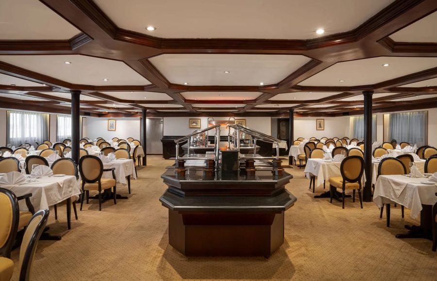 A wide-angle view of the large and elegant dining room on the Odyssey Nile Cruise, showing rows of set tables with white linen and yellow-upholstered chairs surrounding a central dark wood buffet station.