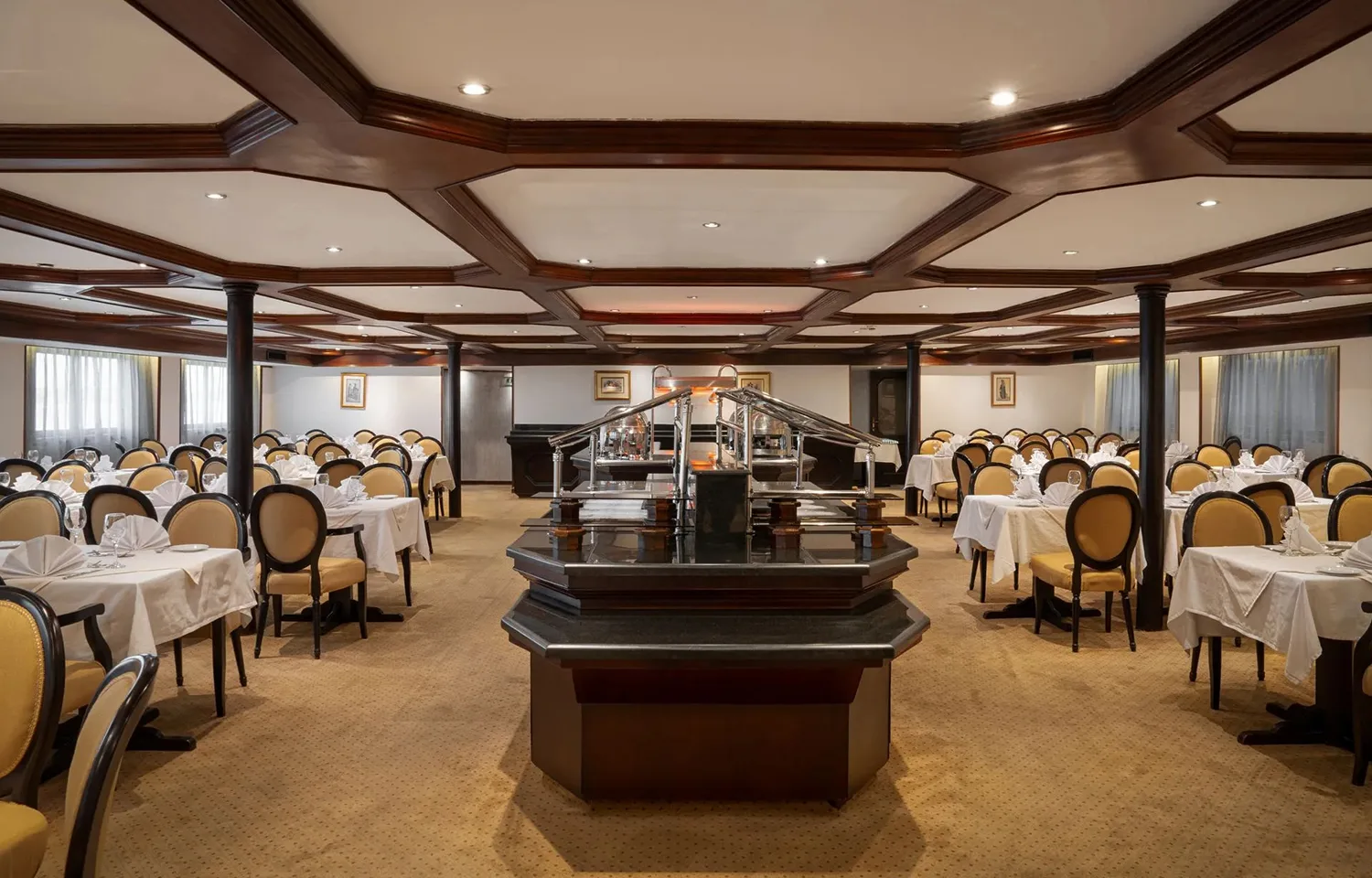 A wide-angle view of the large and elegant dining room on the Odyssey Nile Cruise, showing rows of set tables with white linen and yellow-upholstered chairs surrounding a central dark wood buffet station.