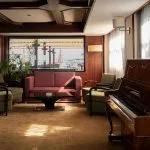 An elegant cruise ship lounge area featuring a wooden upright piano, green armchairs, a red sofa, and a large window providing a view of the outside deck and river.