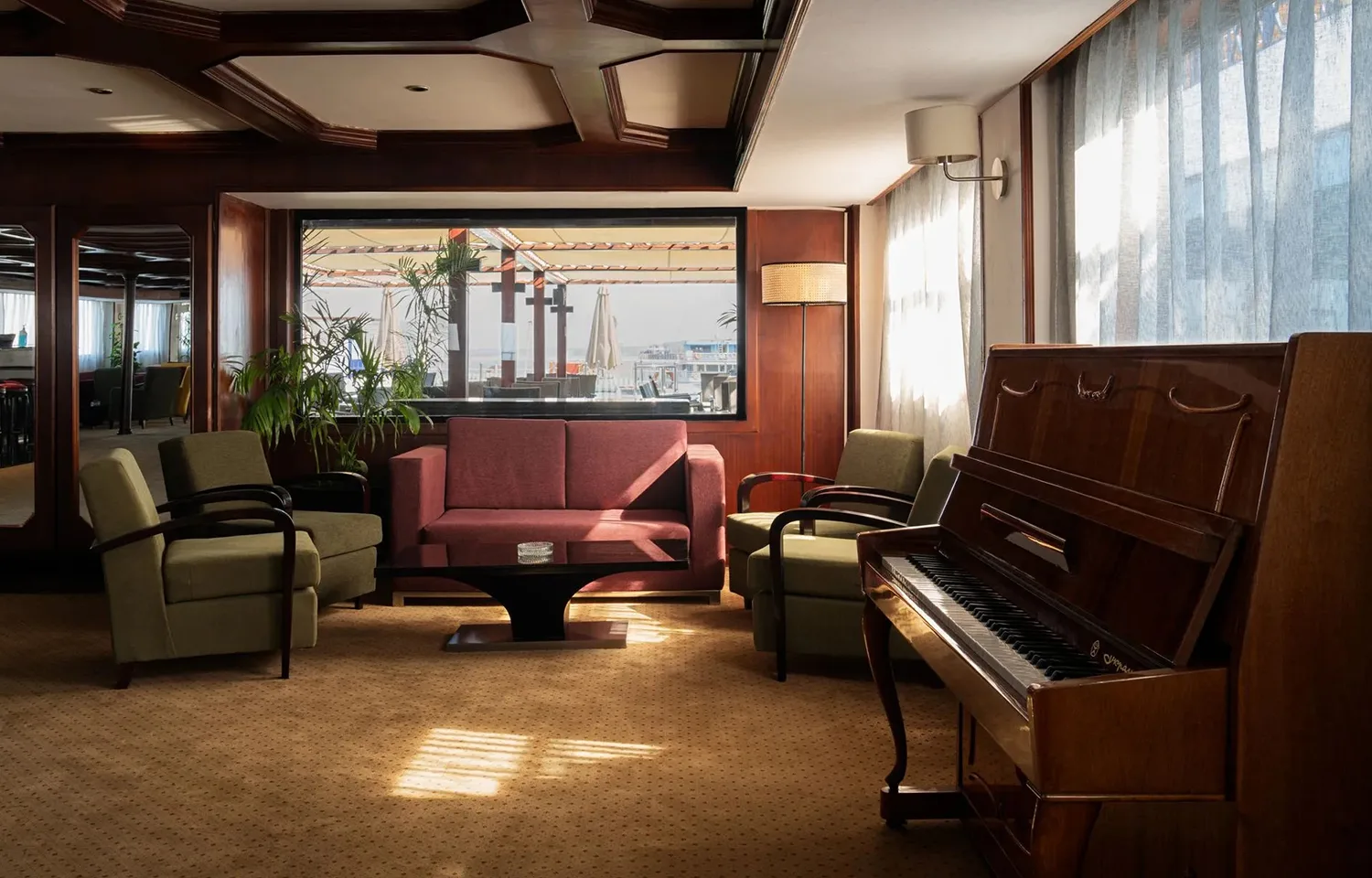 An elegant cruise ship lounge area featuring a wooden upright piano, green armchairs, a red sofa, and a large window providing a view of the outside deck and river.
