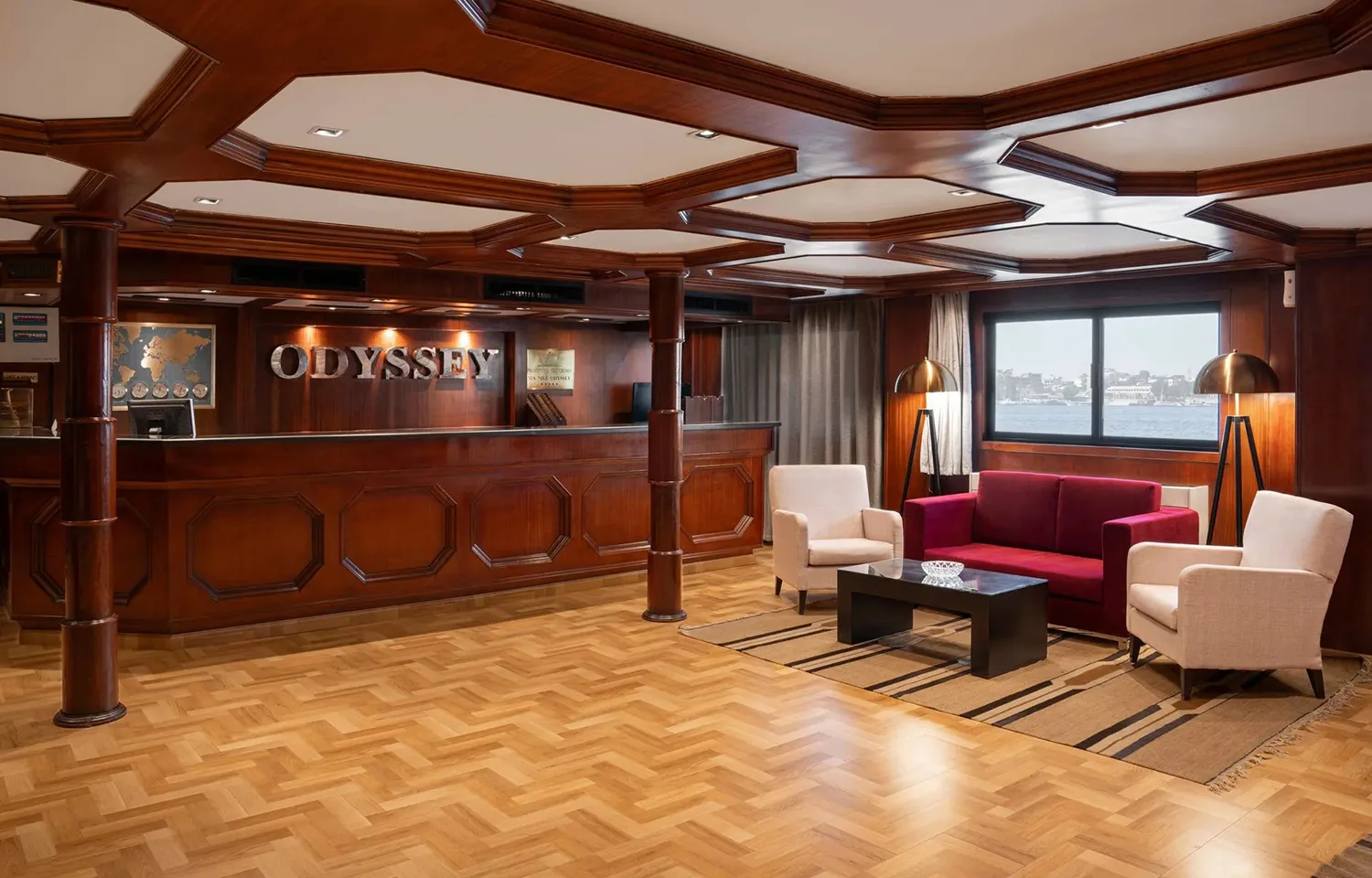 The lobby lounge and reception area of the MS Odyssey, featuring a dark wood reception desk, comfortable armchairs, a red sofa, and a large window overlooking the Nile.