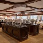 The buffet serving area of the Odyssey Nile Cruise restaurant, showing two dark wood buffet islands topped with multiple large, silver chafing dishes. Dining tables with yellow-upholstered chairs are visible along the windowed walls.
