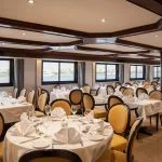 The elegant, spacious dining room of the Odyssey Nile Cruise, featuring multiple round tables covered with white linen tablecloths, set with place settings, and surrounded by mustard-yellow upholstered chairs. Large windows line the side, offering views of the Nile River.