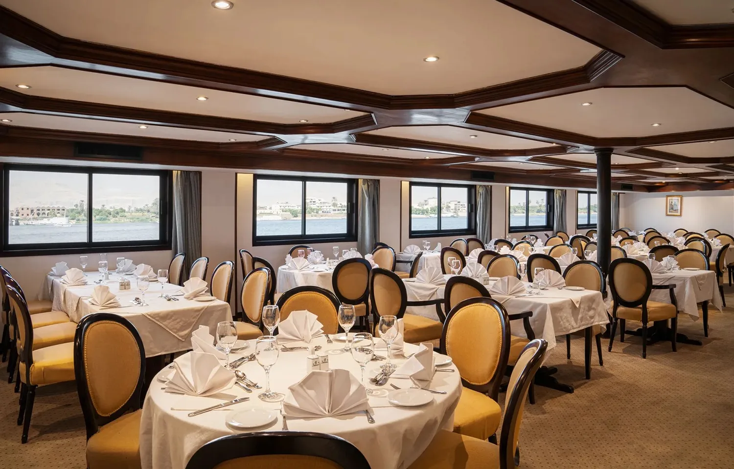 The elegant, spacious dining room of the Odyssey Nile Cruise, featuring multiple round tables covered with white linen tablecloths, set with place settings, and surrounded by mustard-yellow upholstered chairs. Large windows line the side, offering views of the Nile River.