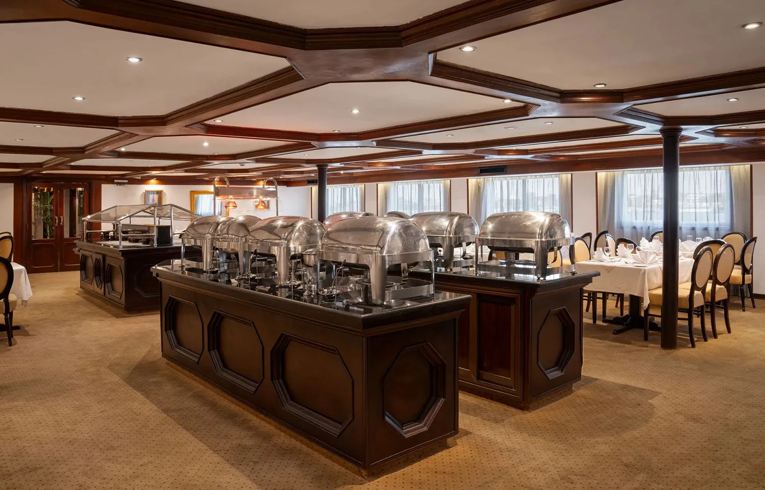 The buffet serving area of the Odyssey Nile Cruise restaurant, showing two dark wood buffet islands topped with multiple large, silver chafing dishes. Dining tables with yellow-upholstered chairs are visible along the windowed walls.