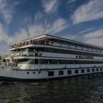 Wide-angle photograph of the MS Nile Odyssey cruise ship sailing on the river under a blue, cloudy sky. The ship is large, with white and green trim, and its name is clearly visible.