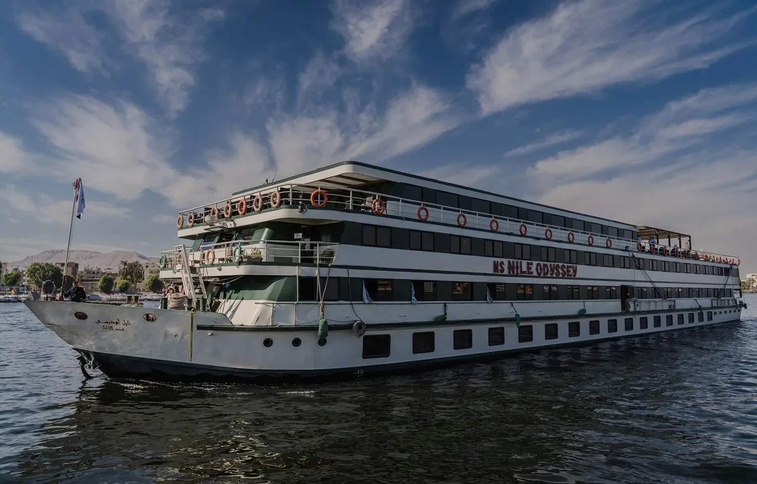 Wide-angle photograph of the MS Nile Odyssey cruise ship sailing on the river under a blue, cloudy sky. The ship is large, with white and green trim, and its name is clearly visible.