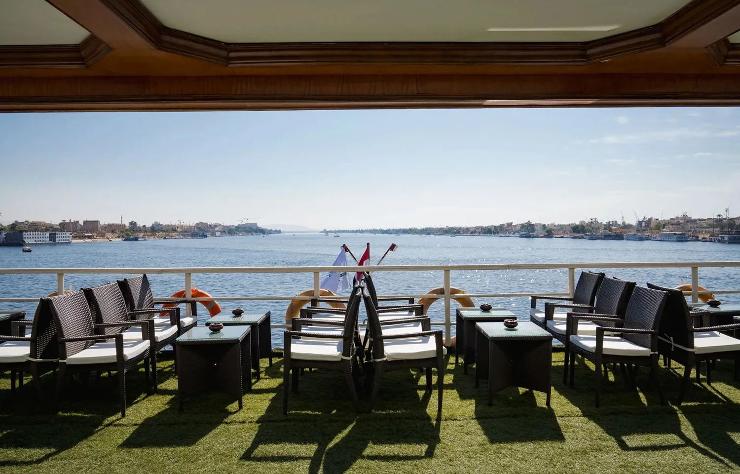 Wicker chairs and tables with white cushions arranged on the sundeck of the Odyssey Nile Cruise, overlooking the wide, blue Nile River with palm trees and a city skyline visible on the distant banks under a clear blue sky.
