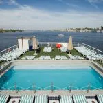 Panoramic view of the swimming pool and sun deck of the MS Odyssey cruise ship, overlooking the wide River Nile under a bright blue sky.