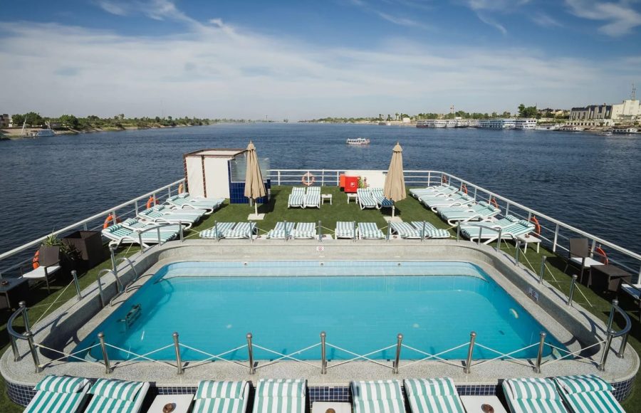 Panoramic view of the swimming pool and sun deck of the MS Odyssey cruise ship, overlooking the wide River Nile under a bright blue sky.