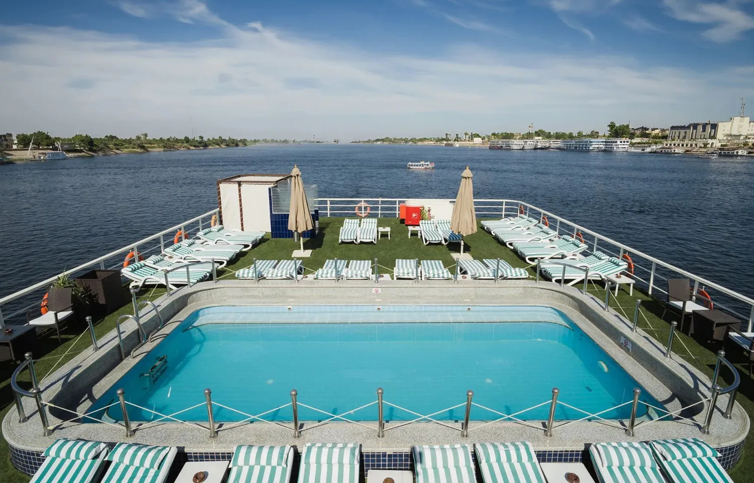 Panoramic view of the swimming pool and sun deck of the MS Odyssey cruise ship, overlooking the wide River Nile under a bright blue sky.