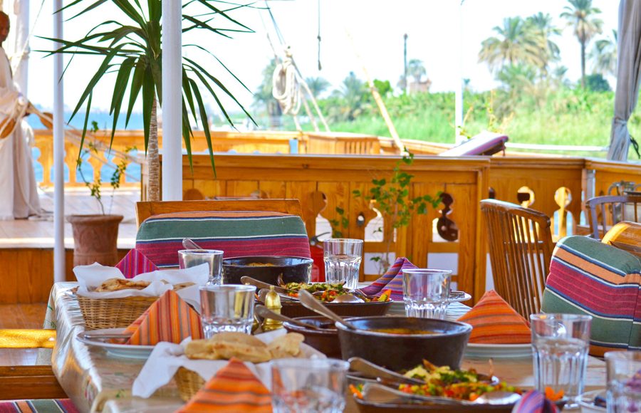 A dining table on a boat deck set with Egyptian dishes, colorful napkins, and bread baskets.