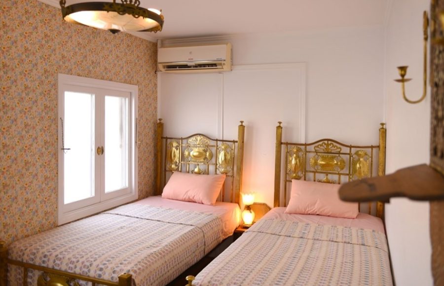 An elegant twin cabin on a Dahabiya Nile cruise featuring brass bed frames, floral wallpaper, and a window view.