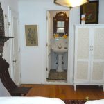 A bright bedroom on a Dahabiya Nile cruise boat featuring white walls, wood floors, an ornate dark wood chair, and a view into a private bathroom with a pedestal sink.