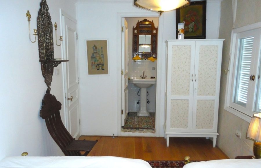 A bright bedroom on a Dahabiya Nile cruise boat featuring white walls, wood floors, an ornate dark wood chair, and a view into a private bathroom with a pedestal sink.