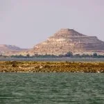 Panoramic view of the vast Salt Lake Aftanas near Siwa Oasis, with its greenish-blue water, a small band of trees on the opposite shore, and distinctive tiered, rocky mountains in the far background under a pale sky.