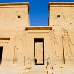 Facade of Philae Temple, Aswan, Egypt. Large stone entrance pylon with carved reliefs of pharaohs and gods under a bright blue sky.