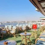 Outdoor deck of a Nile cruise ship with rows of wicker chairs and small tables, overlooking the Nile River and the distant cityscape.