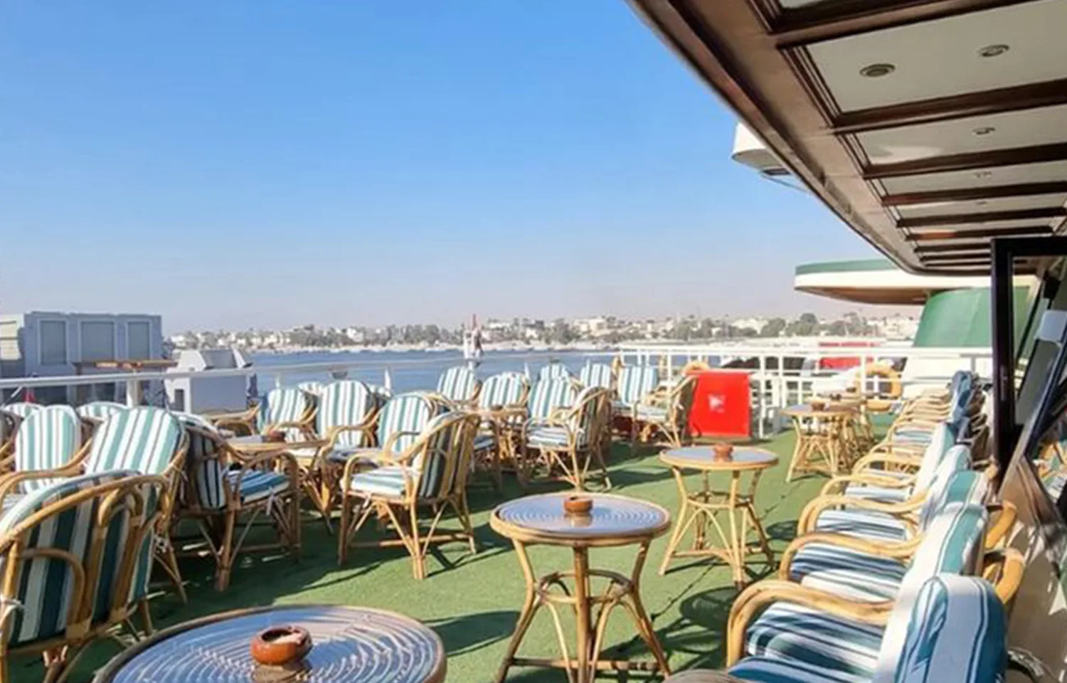 Outdoor deck of a Nile cruise ship with rows of wicker chairs and small tables, overlooking the Nile River and the distant cityscape.