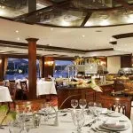 A close-up view of a set dining table in the formal restaurant of a Nile cruise ship, showing a white tablecloth, place settings, wine glasses, and a centerpiece, with a buffet and other tables visible in the background through wooden columns and large windows.