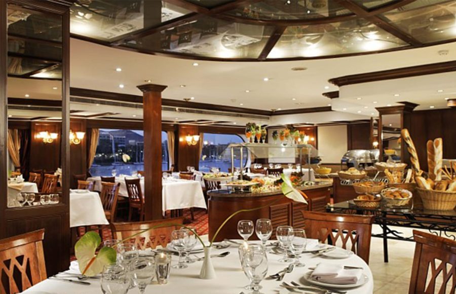 A close-up view of a set dining table in the formal restaurant of a Nile cruise ship, showing a white tablecloth, place settings, wine glasses, and a centerpiece, with a buffet and other tables visible in the background through wooden columns and large windows.