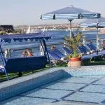The sundeck of a Nile cruise ship featuring a rectangular swimming pool with blue and white tiled edges, blue and white striped umbrellas and loungers, a blue canopy swing, and a view of the Nile River with traditional white sailboats (feluccas) in the background.