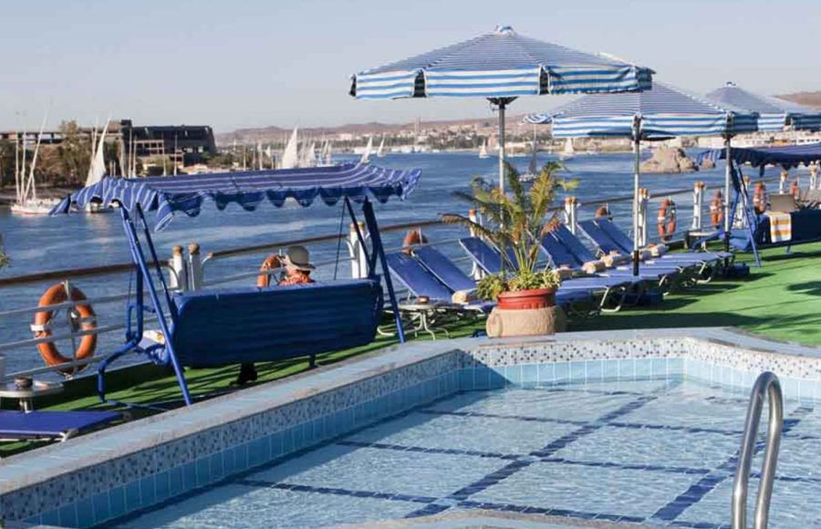 The sundeck of a Nile cruise ship featuring a rectangular swimming pool with blue and white tiled edges, blue and white striped umbrellas and loungers, a blue canopy swing, and a view of the Nile River with traditional white sailboats (feluccas) in the background.