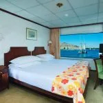 A bright single cabin on a Nile cruise ship featuring a double bed with a colorful orange and yellow floral throw, dark wooden furniture, and a large window offering a view of the river and passing traditional sailboats (feluccas).