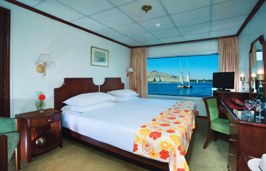 A bright single cabin on a Nile cruise ship featuring a double bed with a colorful orange and yellow floral throw, dark wooden furniture, and a large window offering a view of the river and passing traditional sailboats (feluccas).