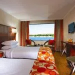 A twin-bedded cruise ship cabin with a large window overlooking a river and lush green banks. The room has twin beds with white linens, orange and white patterned throws, and wooden furniture including a headboard, desk, and side table.