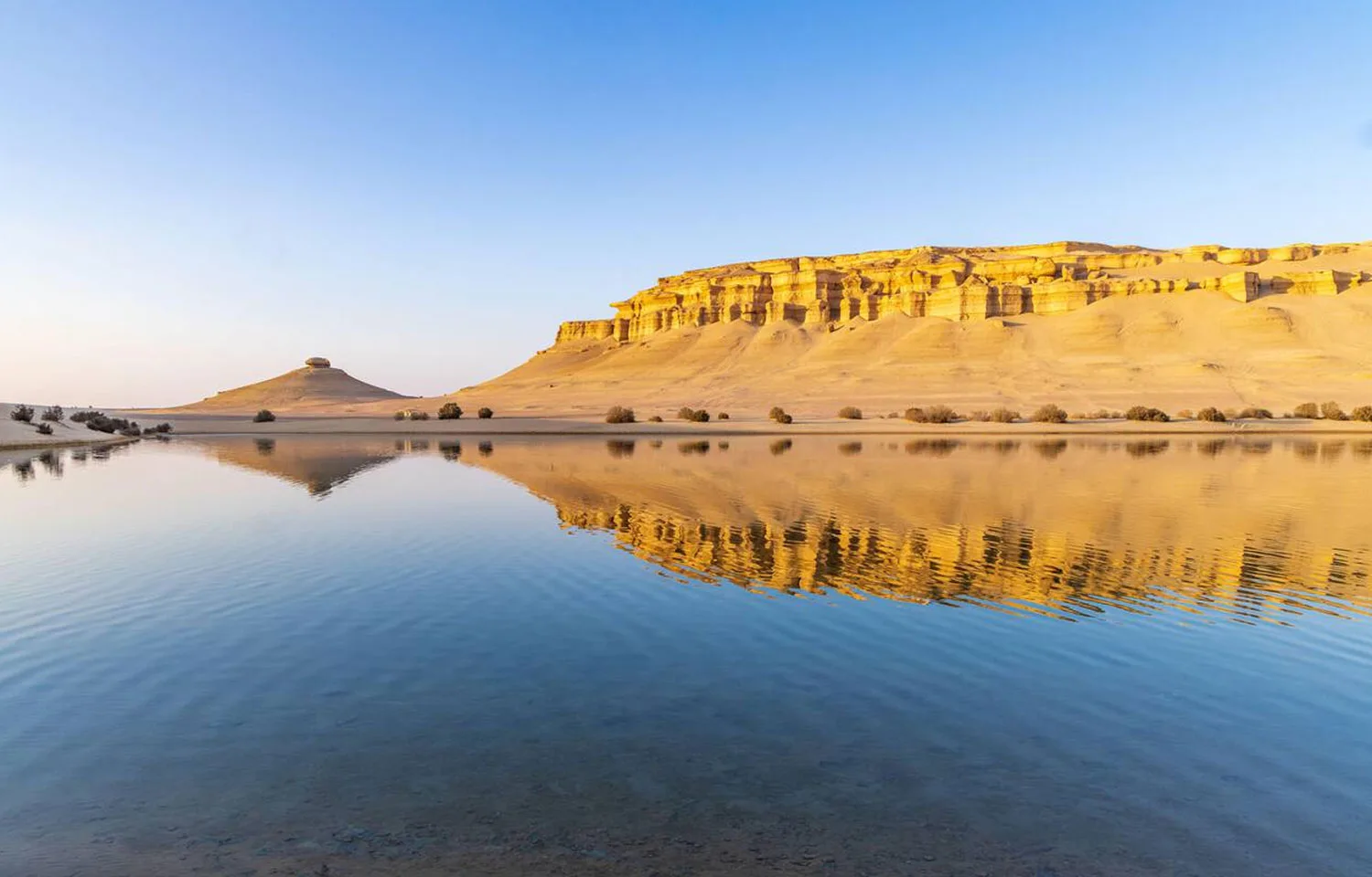 Golden Reflections in Qarun Lake at Fayoum Oasis, showing a calm water surface mirroring the sharply defined desert cliffs and a small conical hill under a clear blue sky.