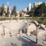 The ruins of the Roman Amphitheatre Alexandria Egypt, showing the white marble seating tiers of the ancient structure, surrounded by green grass and modern city buildings. This stop is key for any Egypt travel itinerary 10 days.